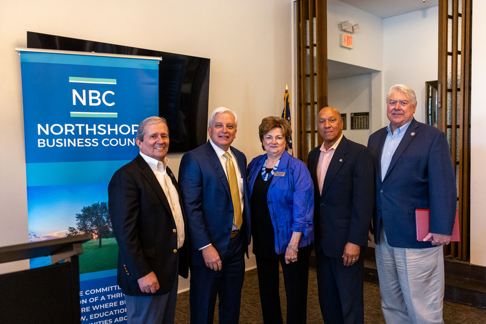 Five business professionals stand together for a photo beside an NBC NorthShore Business Council banner in a conference room. A flag is visible in the background.