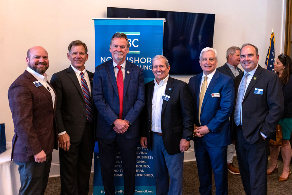 Six men in suits stand in a row posing for a photo at a professional event, with a blue banner and an American flag in the background (text includes NBC North Shore).