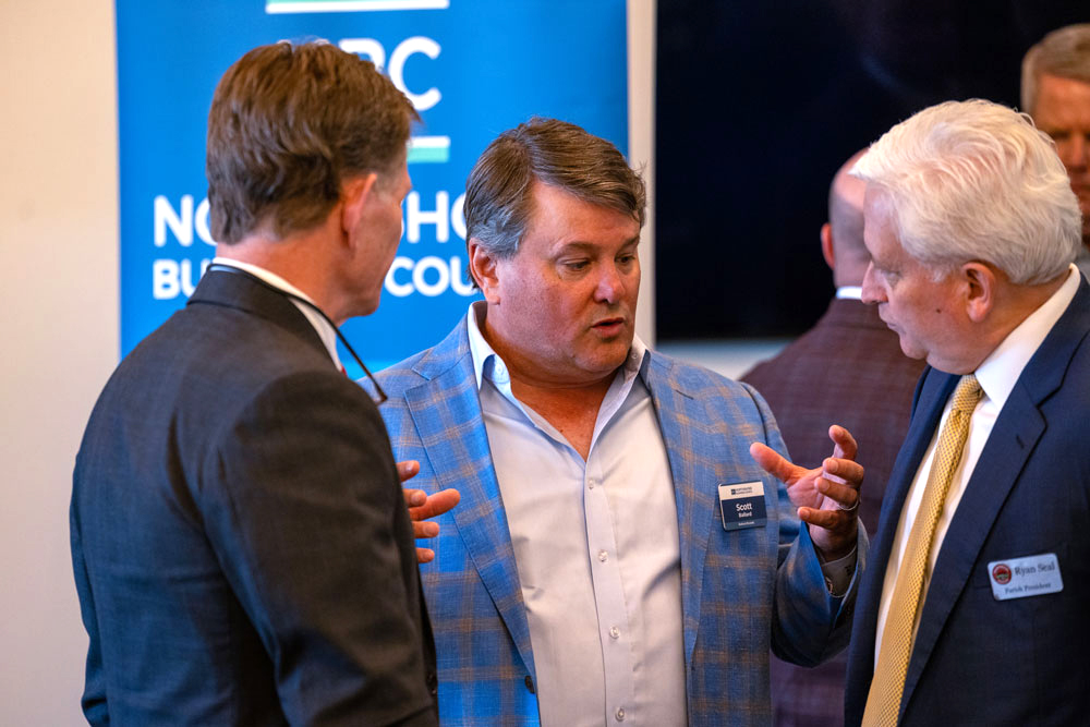 Three men in business attire converse at a networking event, with a blue banner in the background.