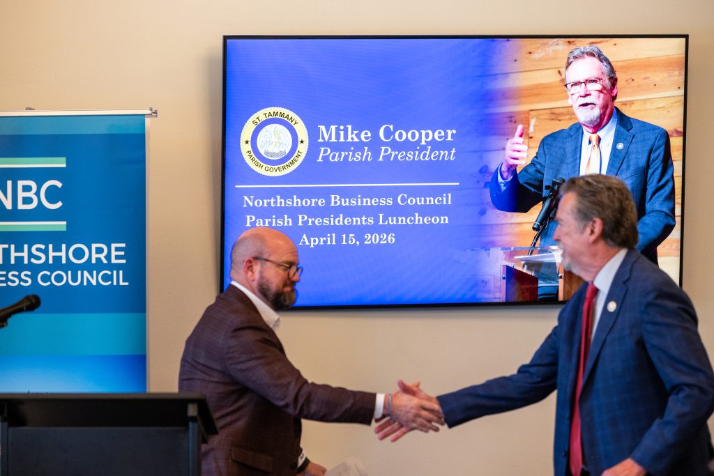Two men in suits shake hands in front of a large screen that reads 'Mike Cooper Parish President' and shows a parish seal, at a formal event.