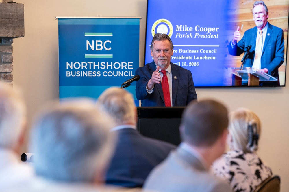 Speaker in a dark suit and red tie at a podium delivering a talk, with an NBC Northshore Business Council banner nearby and a large screen behind him showing another speaker.