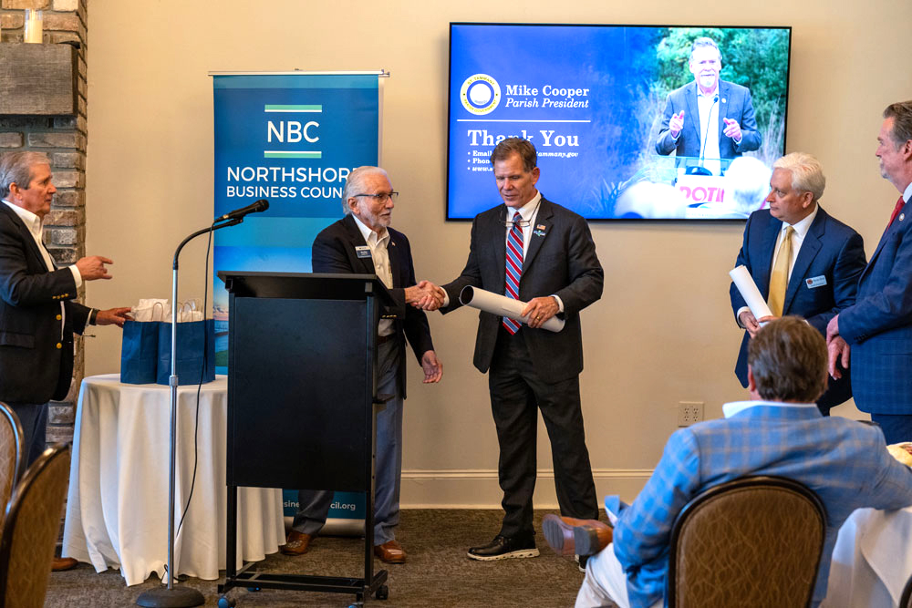 Business event: two men shake hands near a podium as others watch, NBC Northshore banner in the background.