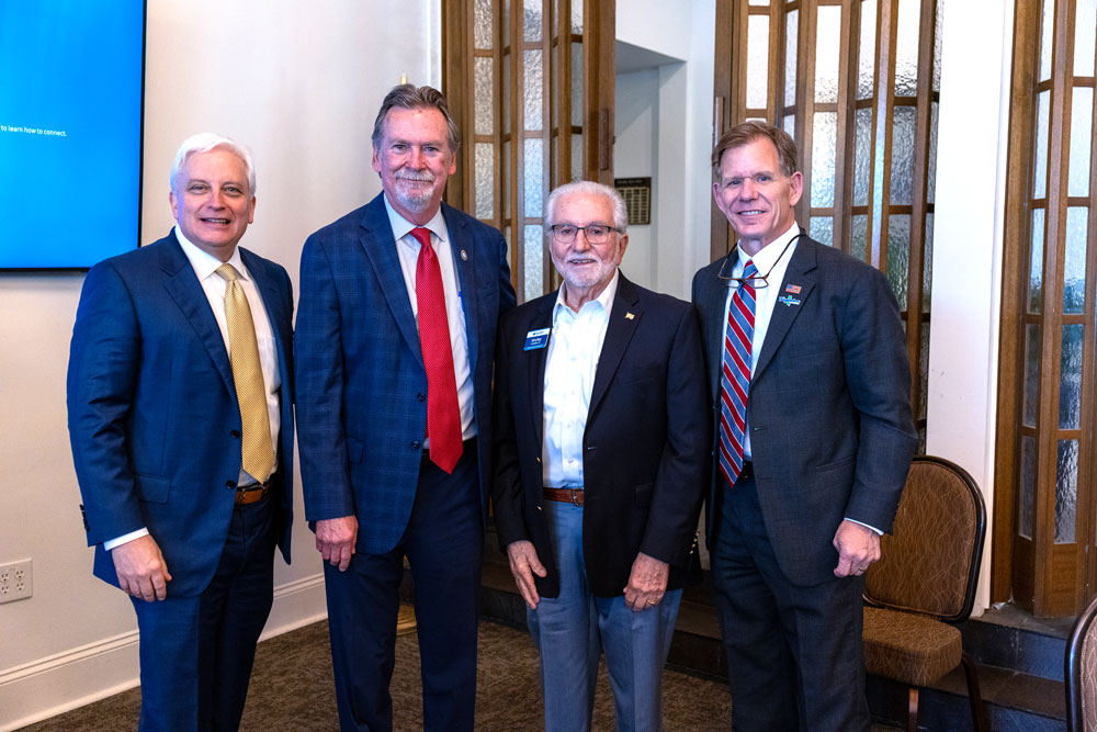 Four men in business suits stand together, posing for a photo at an indoor event, each wearing a name badge. From left: man in blue suit with gold tie, man in blue check suit with red tie, man in dark blazer with white shirt and glasses, man in dark suit with striped tie.