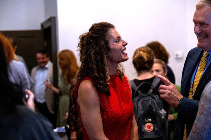 Woman in a red lace dress laughs while talking with a man in a dark suit and yellow tie at a social event.
