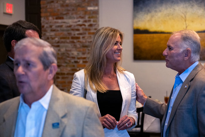 Woman in a white blazer smiles while speaking with a gray-haired man in a suit at a casual reception area.