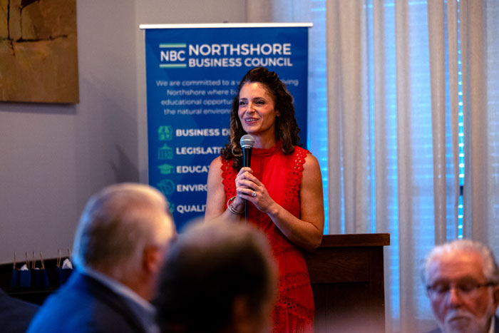 Woman in a red dress speaks into a microphone at a podium during a NBC Northshore Business Council event, with a banner in the background.