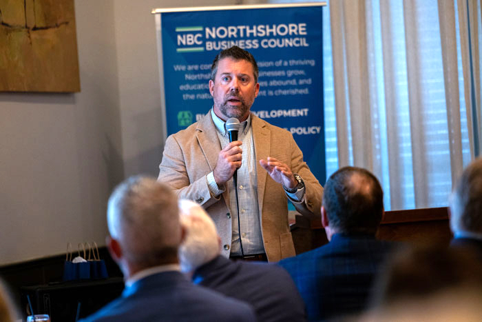Man in a tan blazer speaks into a microphone at a Northshore Business Council event, with a blue banner in the background and attendees nearby.