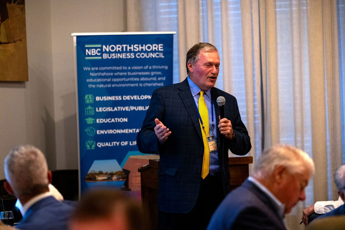 Male speaker in a blue suit and yellow tie addresses a conference from a podium; banner behind reads NBC Northshore Business Council.
