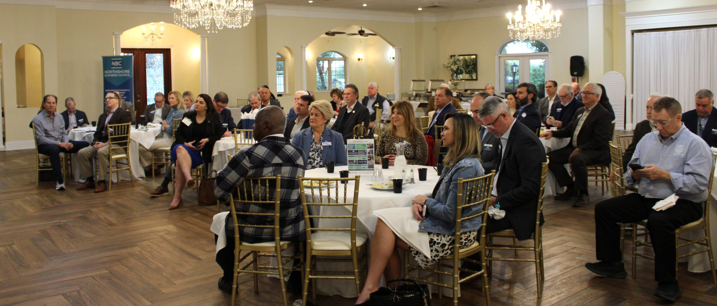 Professionals seated at round tables in a banquet hall during a conference or networking event, with a chandelier overhead and a banner in the background