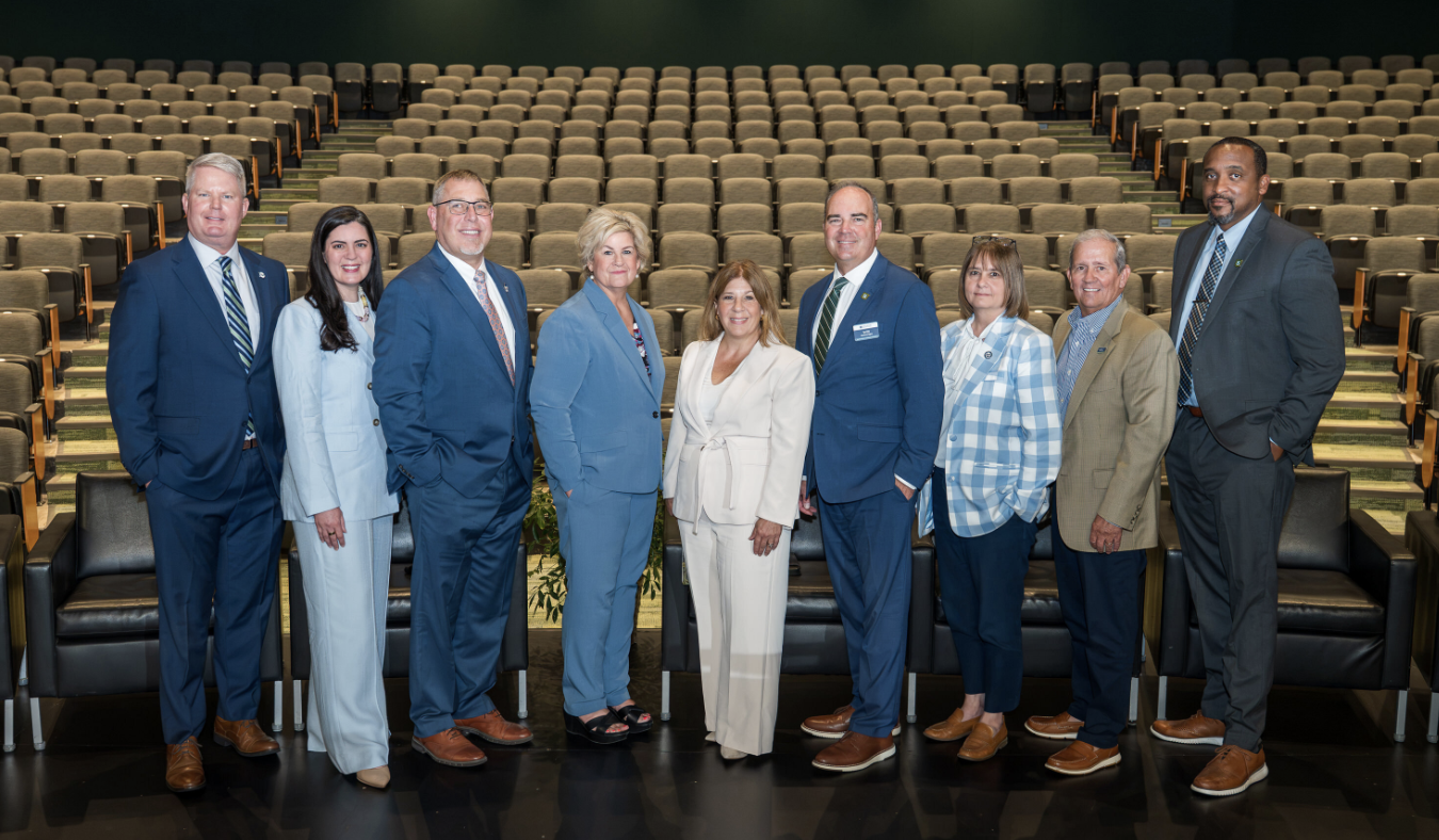 Nine professionals in business attire pose on a stage in an auditorium with empty seats behind them, looking at the camera.